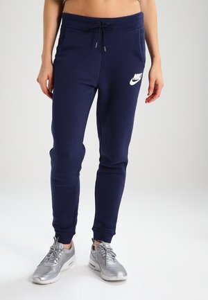 Personne portant un pantalon de survêtement Nike bleu marine avec cordon de serrage et des baskets Nike argentées, debout sur un sol de studio blanc.
