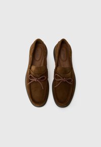 Brown suede shoes featuring a rounded toe, matching lace accent, and a smooth rubber sole. The interior has a tan lining with branding.