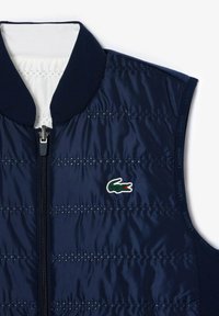 Chaleco acolchado azul marino con un cuello alto, cierre de cremallera y logo de Lacoste. Presenta líneas de costura horizontales y una textura suave y brillante.