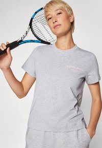 Grå bomullströja med korta ärmar, rundad halsringning och en rosa tryckt logotyp. Modellen håller en svart och blå tennisracket.