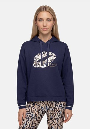 Vrouw met lang blond haar draagt een navy hoodie met een lipgrafiek met luipaardprint en bijpassende luipaardprint broek, staand tegen een effen achtergrond.