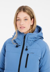Blauwe pufferjack met capuchon, voorzien van een ritssluiting aan de voorkant, elastische trekkoordsluitingen en een klein borstzakje met een zwarte rits.