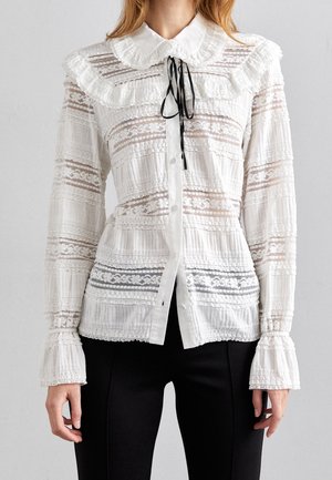 Blusa blanca de encaje de manga larga con cuello y puños con volantes, que presenta un lazo de cinta negra y patrones intrincados de encaje horizontal.