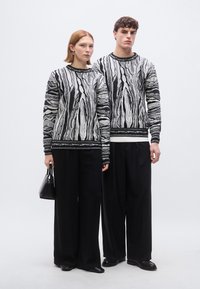 Pulls à motifs noirs et blancs avec un design ondulé, associés à un pantalon noir à jambes larges. Les accessoires comprennent un petit sac noir.