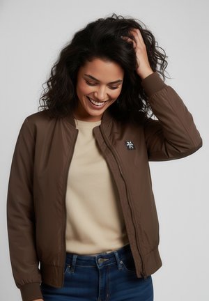 Frau mit lockigem dunklem Haar, die lächelt, trägt eine braune Jacke mit Reißverschluss über einem beigen Hemd und blauen Jeans, berührt mit einer Hand ihr Haar.