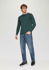Groene gebreide trui met een ronde hals, lange mouwen en geribbelde manchetten, gecombineerd met vervaagde blauwe jeans en zwarte sneakers.