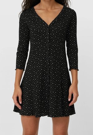 Femme portant une robe noire avec des motifs d'étoiles blanches, boutonnage devant, manches trois-quarts et ourlet évasé.