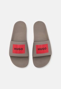 Grijze rubberen sandalen met een gestructureerde voetbed, voorzien van brede rode bandjes die het zwarte "HUGO" logo prominent tonen. Plat ontwerp, casual pasvorm.