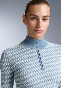 Ljusblå stickad tröja med hög krage och kvartzip, med ett texturerat vågmönster i vitt, tighta ärmar och mjukt tyg.