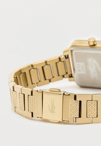 Kultaiset metallikellot, joissa on suorakulmainen kasvo ja teksturoidut ranneketjut. Varustettu kaiverretulla logolla soljessa ja sileällä, kiiltävällä pinnalla.