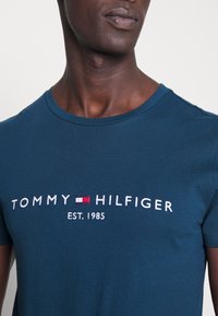 Närbild av en person som bär en blå T-shirt med Tommy Hilfiger-logotypen och "EST. 1985" broderat på bröstet.