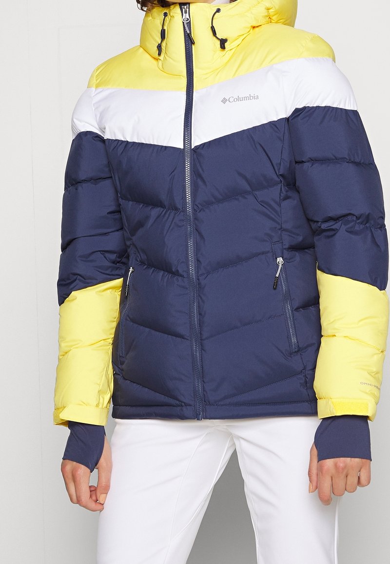 Giacca invernale Columbia con un design a blocchi di colore in blu navy, giallo e bianco, dotata di un colletto alto e tasche laterali con zip.