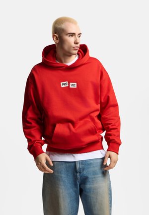 Młody mężczyzna z krótkimi blond włosami, ubrany w czerwoną bluzę z kapturem z napisem "PROSTO." oraz luźne niebieskie jeansy, patrzy w bok na białym tle.