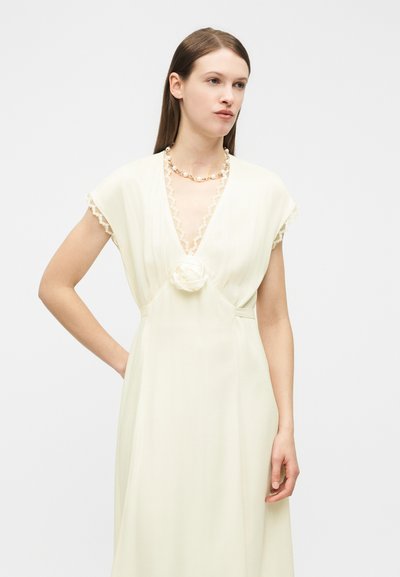 Femme aux cheveux bruns et raides, portant une robe crème avec une bordure en dentelle, un détail florale à la taille et un collier de perles, debout avec un bras plié.