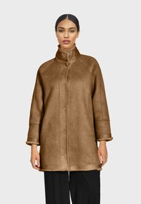 Manteau en fausse fourrure marron avec un col montant, des manches longues et des poches latérales. Il présente une coupe droite et des textures avec une finition douce.