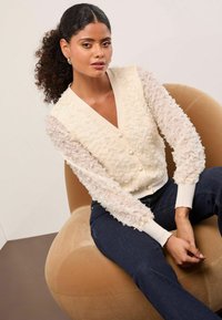 Krémový kardigan s volánovými průhlednými rukávy, zapínáním na knoflíky a žebrovanými manžetami, zkombinovaný s tmavými denimovými kalhotami, usazený na zaobleném béžovém sedadle.