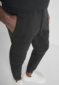 Zwarte joggingsbroek met een gladde textuur, elastische tailleband en zakken. Kenmerkt ribgebreide accenten op de knieën. Gecombineerd met witte sneakers.