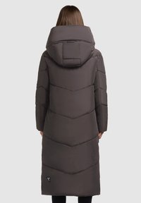 Vrouw in een lange, donkerbruine gewatteerde winterjas met grote kap, staand met haar rug naar de camera tegen een effen, lichte achtergrond.