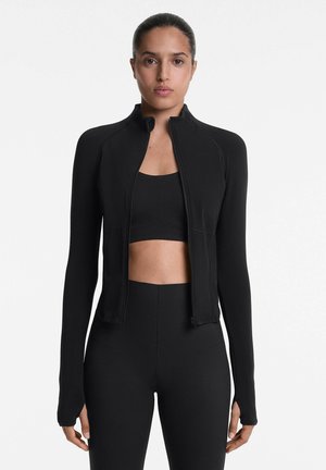 Schwarze Sportjacke mit Reißverschluss und langen Ärmeln, in einer taillierten Form und mit glatter Textur, kombiniert mit einem schwarzen Cropped-Top und Leggings.