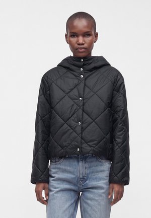 Person iført en sort quiltet dunjakke med trykknapper og lyseblå denimjeans, stående mod en ensfarvet hvid baggrund.
