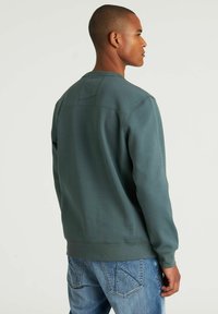 Sudadera verde con escote redondo y puños acanalados, que presenta un panel trasero texturizado, combinada con jeans de mezclilla azul claro.