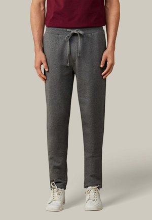 Hombre vestido con pantalones deportivos gris oscuro con cordón y zapatillas blancas, de pie frente a un fondo claro y liso.