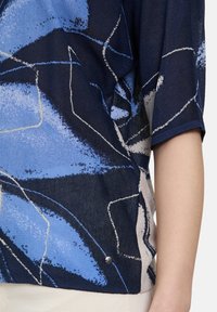 Niebieska i granatowa bluzka w wzory o przezroczystej teksturze. Posiada geometryczne wzory oraz dekoracyjny srebrny guzik przy bocznym szwie.