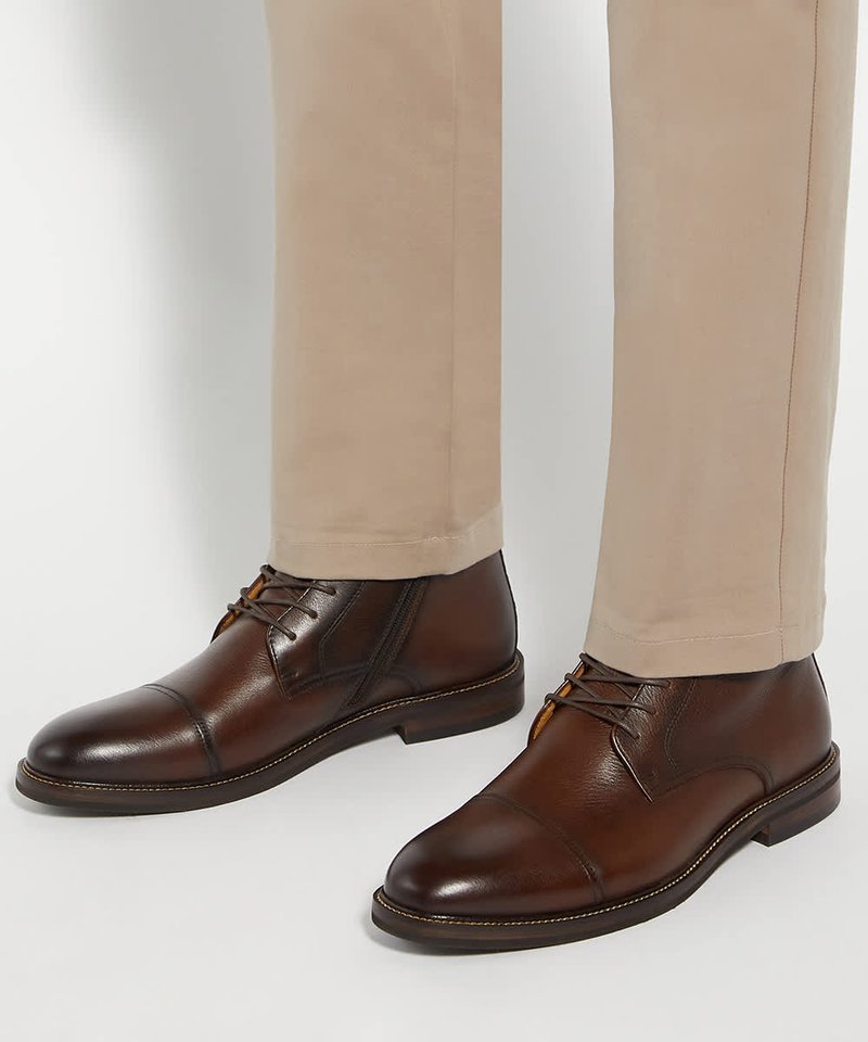 Bottes de ville en cuir marron avec lacets portées avec un pantalon beige sur fond blanc.