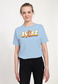 Světle modré bavlněné tričko s grafikou medvěda na kolečkových bruslích a čísly "1982" v tučných, barevných písmenech.