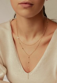 Goldfarbene, geschichtete Halskette mit zarten Ketten unterschiedlicher Längen, die über Textur-Elemente und einen Lariat-Stil in einer neutralen Umgebung verfügt.