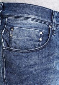 Denim jeans with a faded blue finish, featuring a small front pocket with three metallic rivets and contrasting stitching.