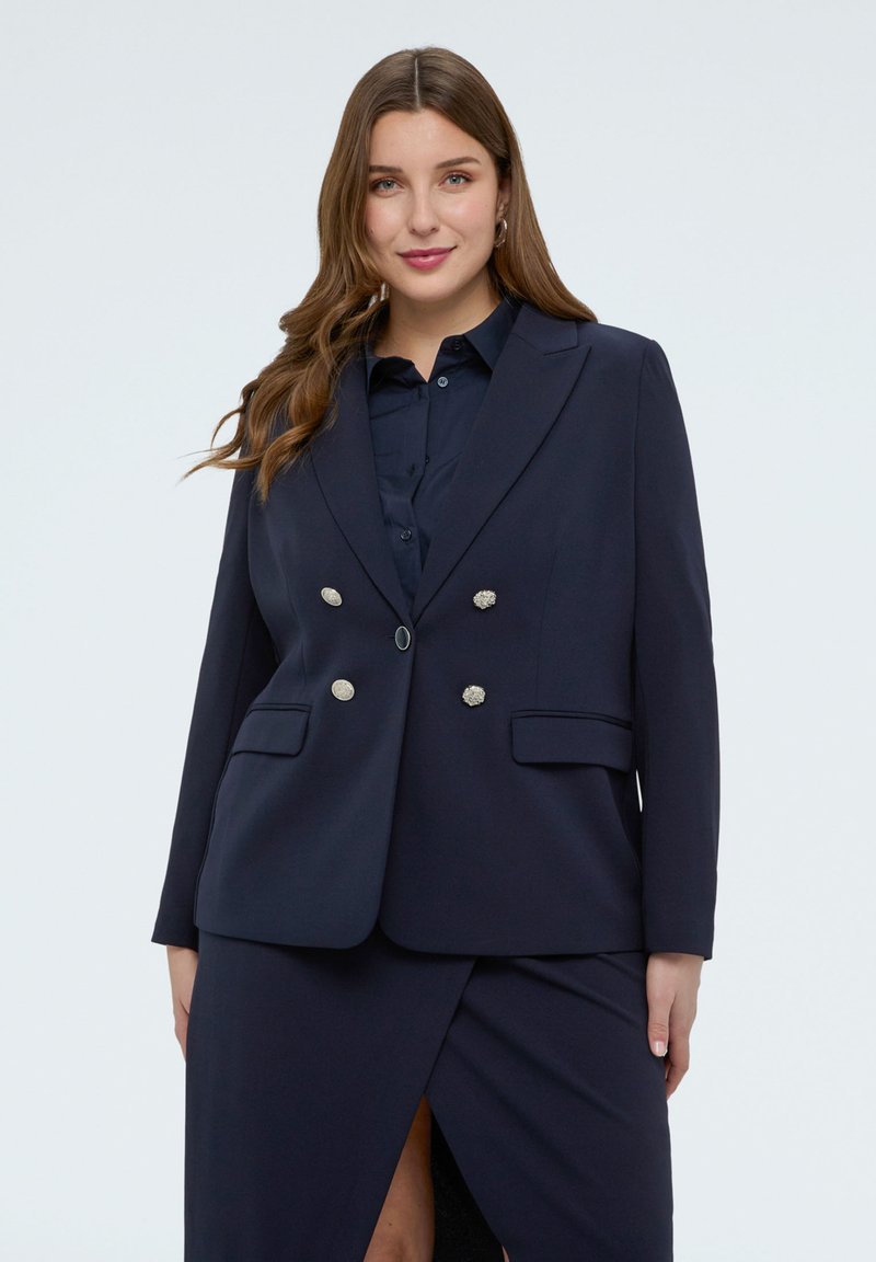 Frau trägt ein marineblaues, doppelt geknöpftes Blazer mit silbernen Knöpfen über einem passenden Hemd und einem marineblauen Rock mit Frontschlitz.