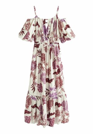 Femme portant une robe blanche à épaules dénudées à motifs floraux dans des tons roses et bordeaux, debout à l'extérieur près d'un mur en pierre avec l'océan en arrière-plan.
