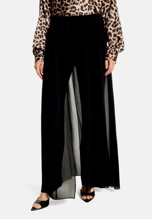 Kvinde iført sorte bukser med vidde ben og transparent overlag, kombineret med sorte åbne hæle og en langærmet bluse med leopardprint.