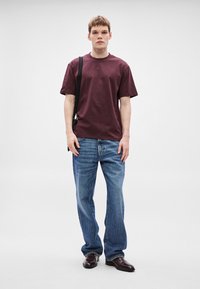 Burgundska majica s kratkimi rokavi v kombinaciji s svetlo modrimi jeans hlačami in rjavimi usnjenimi čevlji; model stoji pokončno, drži črno torbico z nastavljivim trakom.