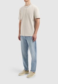 Lichtbeige T-shirt met korte mouwen, ontspannen pasvorm, gecombineerd met lichtblauwe jeans en beige instappers. Subtiele branding op het shirt.