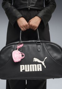 Czarna skórzana torba podróżna z białym logo "PUMA", o zaokrąglonym kształcie i różowym breloczku do kluczy. Gładka tekstura, solidne uchwyty i metalowe akcenty.