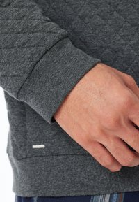 Grijze gewatteerde sweatshirt met een gestructureerde stof, ribbelmanchetten en een kleine witte accent bij de zoom. Focus op de hand bij de zak.
