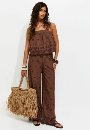Femme portant un haut sans manches brodé marron et un pantalon large assorti, tenant un grand sac en paille marron à franges, portant des bracelets et des sandales.