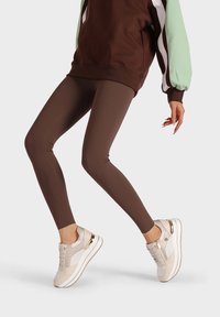 Leggings marroni abbinati a un felpa marrone e verde menta, e sneakers bianche con dettagli dorati. I leggings sono aderenti e di una consistenza liscia.