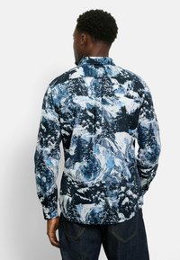 Langarmhemd mit einem blauen und weißen Druck von Bergen und Wäldern. Mit einem Button-Down-Kragen und einem geraden Saum. Der Stoff wirkt glatt.