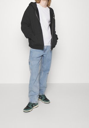 Sudadera negra con cremallera y bolsillos laterales, combinada con una camiseta blanca, vaqueros holgados de color azul claro y zapatillas verdes y negras.