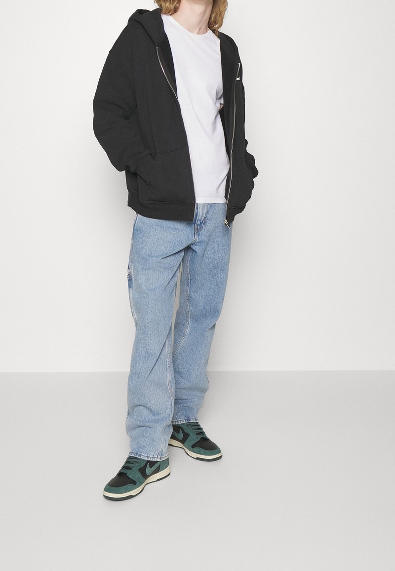 Sudadera negra con cremallera y bolsillos laterales, combinada con una camiseta blanca, vaqueros holgados de color azul claro y zapatillas verdes y negras.