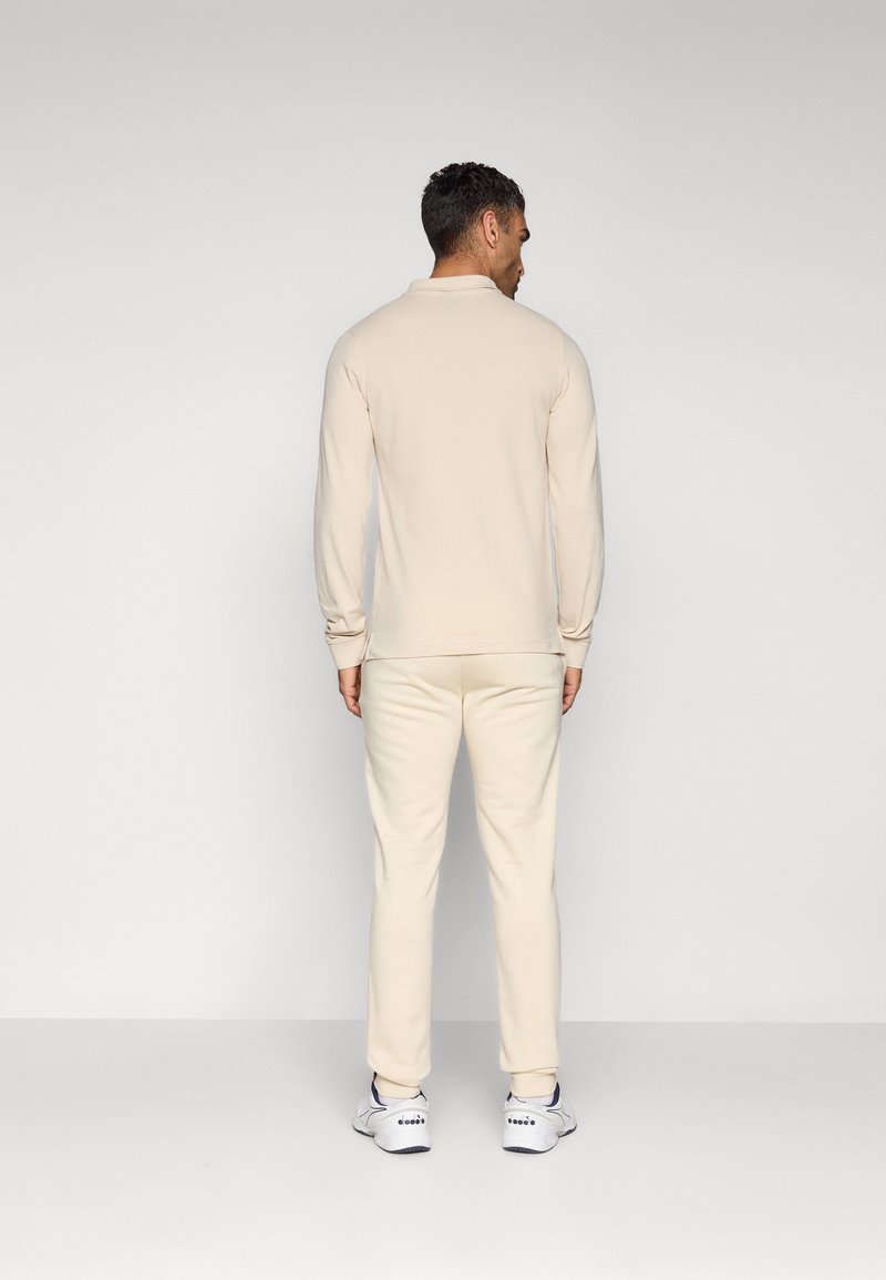 Mann in beigem Langarmhemd und passender Hose, der mit dem Rücken zu einem schlichten hellgrauen Hintergrund steht.