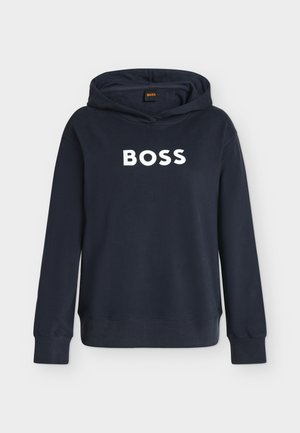 Femme aux cheveux blonds portant un sweat à capuche bleu marine avec le texte "BOSS", un pantalon large marron, des baskets blanches et tenant un petit sac à main blanc.