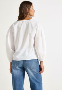 Camicia bianca a maniche lunghe con texture, maniche a sbuffo, scollo rotondo e polsini elastici. Abbinata a pantaloni oversized di jeans blu chiaro.