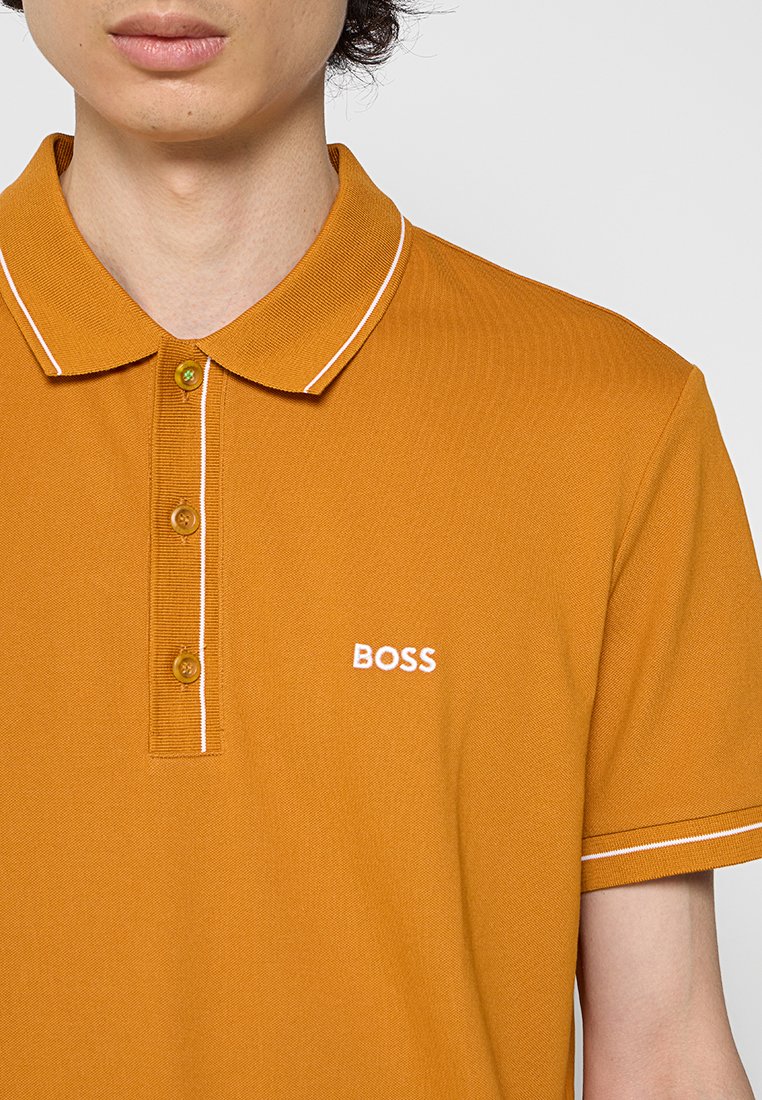Polo di colore giallo senape con tessuto testurizzato, placca con tre bottoni, rifinitura del colletto bianca e un piccolo logo "BOSS" bianco sul petto.