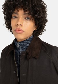 Veste en coton brun avec un col en velours côtelé, portée sur un pull à col montant texturé navy. Gros plan sur le visage avec un maquillage naturel et des cheveux bouclés.