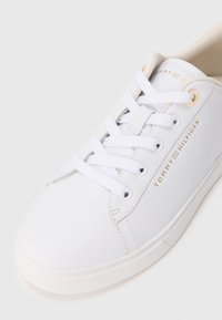 Hvit Tommy Hilfiger sneakers med hvite snørebånd, gulløyler og gull logo tekst på siden og tungen, vist ovenfra på hvit bakgrunn.