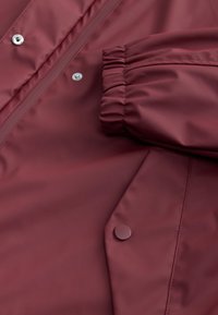 Section de veste imperméable bordeaux montrant des boutons pression, une fermeture éclair, un poignet élastique et une poche à rabat inclinée avec bouton.